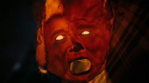 Close-up of a lampshade crudely stitched together using the skin from someone’s head — including dark curly hair, eye holes, nose, and a gaping mouth. On the right side of the image, ASL performer Justin Jackerson signs “mask/expression.” 