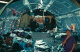 Inside a makeshift shelter with metallic walls, numerous women lie on the ground covered in reflective emergency blankets. In the midground, a long folding table holds an orange water cooler and various supplies. In the background, Deandra reaches out her hand to someone lying on the ground. ASL interpreter Otis Jones signs “speaking.”