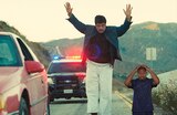 Sensei Sergio St. Carlos stands on the side of a mountain road with his hands raised in surrender. A police patrol car with flashing lights is parked behind him. ASL interpreter Otis Jones signs “hands in the air.”