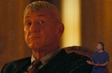Close-up of Col. Lockjaw wearing a suit and tie and sitting in a chair in a dimly lit room. ASL interpreter Otis Jones signs “propose.”