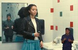 Willa stands in a tiled restroom holding a slim black and silver device in her right hand. Deandra is standing out of frame, but her reflection can be seen in the mirror behind Willa. ASL interpreter Otis Jones signs “calm.”