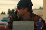 Close-up of Bob Ferguson talking into the receiver of a pay phone while looking to his right. He is wearing a knit hat, wraparound sunglasses, and a red plaid bathrobe. ASL interpreter Otis Jones signs “hey.”