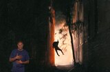 A narrow alleyway at night is illuminated by fire or explosion, with a figure mid-air, seemingly thrown by a blast. ASL interpreter Otis Jones signs “breathing.”