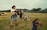In an open grassy field, Willa holds a large firearm ready to fire. Behind her, a group of four women in plain, modest dresses and white head coverings sit in folding chairs. And to her left, a woman wearing a black dress and a white head covering peers through the scope of a large firearm set up on a wooden makeshift perch. ASL interpreter Otis Jones signs “shooting.”