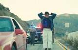 Sensei Sergio St. Carlos stands on the side of a mountain road with his hands on his head as two police officers approach him from behind. Their patrol car with flashing lights is parked behind them.