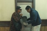 Bob Ferguson and Sensei Sergio St. Carlos stand in a hallway in front of a closed door. Bob, wearing a knit hat, wraparound sunglasses, and a red plaid bathrobe, has his hands clasped together. Sergio St. Carlos bows slightly and offers Bob a battered violin case.