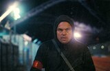 Bob Ferguson, wearing a black hooded sweatshirt and a red armband, walks along a dimly lit industrial area at night with bright lights and fencing in the background.
