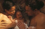 Perfidia and Bob lie in bed cradling a crying newborn baby between them.