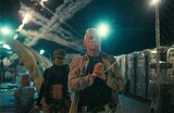 Perfidia and Col. Lockjaw walk through a gated area at night lined with portable toilets. Streaks of smoke line the sky. Lockjaw stands in front; his hands are bound in front of him with a zip tie. Perfidia walks behind him and holds a gun pointed at his back.