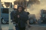 Perfidia wears a hooded jacket and runs away from a car accident. Two smashed vehicles and a hooded figure are behind her.