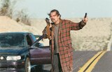 Bob stands in the middle of a road in the desert holding a rifle in his right hand and a small, black device in his left hand. A dark purple sedan is parked behind him with the driver’s side door open.