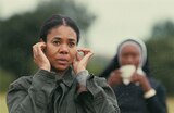 Deandra stands outdoors with her fingers in her ears to block out sound. A blurry figure in a nun’s habit stands behind her, holding a white object to their mouth.