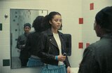 Willa and Deandra stand in a tiled restroom. Willa faces the camera and holds a slim black and silver device in her right hand. Deandra’s back is to the camera, but her face is seen in the mirror behind Willa.