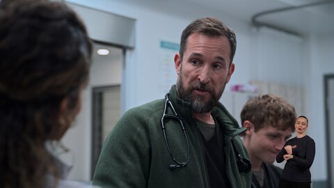 Dr. Robby looks at someone who has their back to the camera. Behind him, Dennis Whitaker looks down at something out of frame. On the right side of the image, ASL performer Amelia Hensley signs "doctor.”