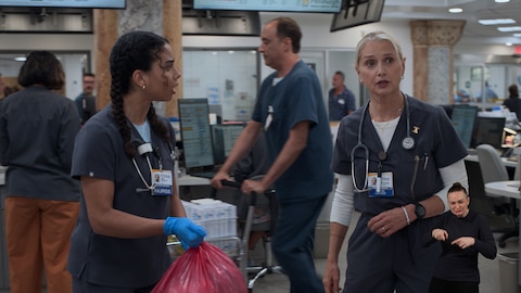 Nurse Emma wears gloves and holds a large red biohazard bag while looking at Nurse Dana Evans, who looks at something out of frame. In the background, other staff members move through the area. On the right side of the image, ASL performer Amelia Hensley signs "pointing to.” 