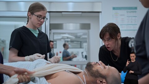 Dr. Mel King and Dr. Cassie McKay are in a hospital treatment room with a male patient lying on a stretcher. The patient has monitoring wires attached to his chest and a breathing tube in his nose. On the right side of the image, ASL performer Amelia Hensley signs "two.” 