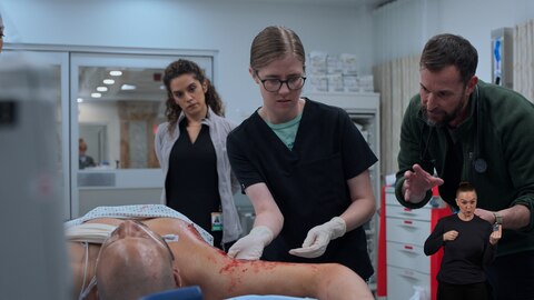 In a hospital treatment room, a patient lies on a stretcher with a bloodied arm exposed. Dr. Mel King wears gloves while examining the arm. Dr. Robby stands to Dr. King's left, giving instructions. And Dr. Baran Al-Hashimi stands behind them, observing. On the right side of the image, ASL performer Amelia Hensley signs "do this” or "hold." 
