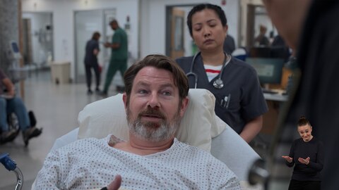 A patient wearing a hospital gown lies on a gurney in a busy hospital corridor. Nurse Princess Dela Cruz stands behind the patient, while another person is partially visible in the foreground. On the right side of the image, ASL performer Amelia Hensley signs "suggest."
