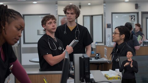 Dennis Whitaker stands at a hospital workstation, holding a folding clipboard and looking up at something out of frame. James and Joy look at Dennis, while Dr. Parker Ellis looks down at a computer monitor. On the right side of the image, ASL performer Amelia Hensley signs "read" or "meaning."
