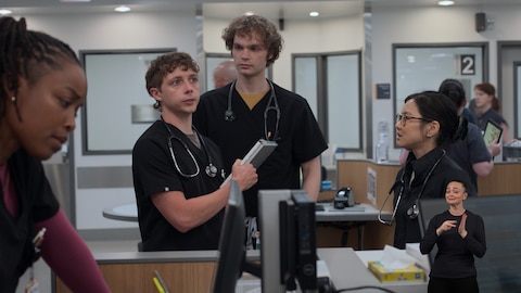 Dennis Whitaker stands at a hospital workstation, holding a folding clipboard and looking up at something out of frame. James and Joy look at Dennis, while Dr. Parker Ellis looks down at a computer monitor. On the right side of the image, ASL performer Amelia Hensley signs "read" or "meaning." 