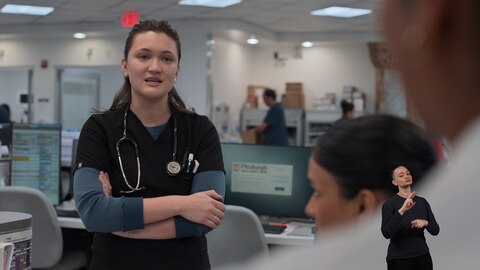 Dr. Trinity Santos stands with her arms crossed amid hospital workstations. In the middle ground, Victoria Javadi looks up at her, while another person is partially visible in the foreground. On the right side of the image, ASL performer Amelia Hensley signs "Rx." 