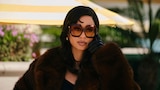 Maddy sits outdoors on a sunny day, wearing oversized square sunglasses, a dark fur coat, and gloves. She holds a cell phone to her face.