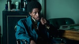 Bishop sits in a chair beside a poker table with playing cards on it. He wears a lightweight jacket and holds the magazine of a handgun close to his face.