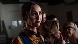 Lexi stands in the foreground wearing a patterned shirt and a dark vest, with headphones around her neck. Several people stand close behind her, also wearing headsets.