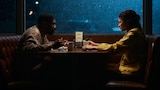 Ali and Rue sit across from each other in a diner booth at night. Ali leans over his plate of food while Rue leans back. A large window in the background is streaked with rain.