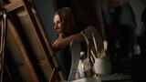 Jules leans forward toward a canvas in a dimly lit studio. Paintbrushes and art supplies are in the foreground.