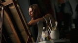 Jules leans forward toward a canvas in a dimly lit studio. Paintbrushes and art supplies are in the foreground.