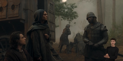 In a foggy forest clearing, King Aegon II wears a hooded robe and stands facing an armored soldier; Larys Strong sits or kneels beside him. A large wooden caravan is parked in the middle ground, the backdoor open, and stairs down. In the background, three armored soldiers focus on something on the ground. In the bottom right-hand corner, ASL performer Landen Gonzalez signs “Sign.”