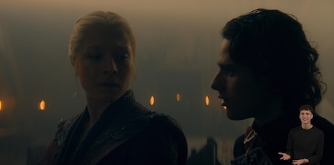 Rhaenyra Targaryen and her son Jacaerys Velaryon stand side by side in a dim, candlelit interior. Rhaenyra has a slightly concerned expression while Jacaerys looks straight ahead. In the bottom right-hand corner, ASL performer Landen Gonzalez signs “Huh? (disappointed)”.