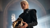 Rhaenyra Targaryen stands in front of a large window with a stone arch. She looks down at a gold crown that she holds with both hands.