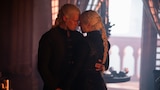 Daemon Targaryen and Rhaenyra Targaryen stand close together inside a dimly lit stone chamber, touching foreheads, holding hands, and staring into each other’s eyes.
