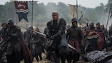A group of wide-eyed soldiers carrying swords, shields, and banners marches across a muddy battlefield led by Daemon Targaryen. Daemon is without a helmet; his hair and face are streaked with blood, and he holds a blood-covered sword at the ready.