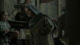 King Aegon II Targaryen stands in the foreground, wearing a headscarf and robe. He holds his left arm at his side, as though it is in a sling. In the background, four men stare at the disgraced king as they eat off their plates with their hands.