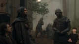 In a foggy forest clearing, King Aegon II wears a hooded robe and stands facing an armored soldier; Larys Strong sits or kneels beside him. A large wooden caravan is parked in the middle ground with the backdoor open and the stairs down. In the background, three armored soldiers focus on something on the ground. In the bottom right side of the image, ASL dubber Landen Gonzales signs “Read.”